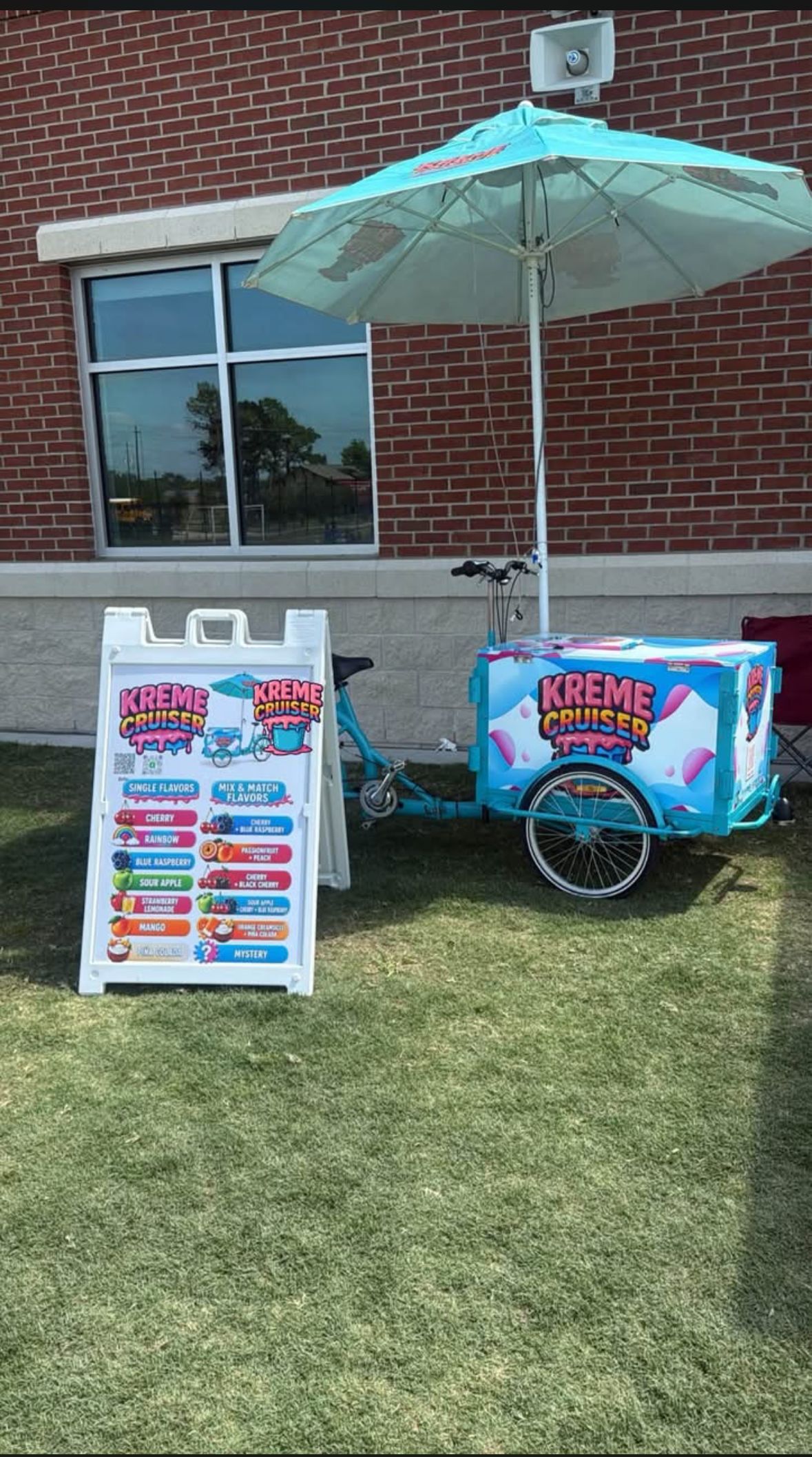 The Kreme Cruiser water ice cart set up on a school lawn under a teal umbrella next to its flavor menu board