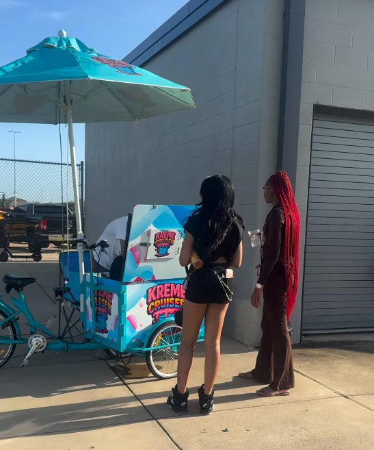 Two guests standing at the Kreme Cruiser cart under its umbrella at an outdoor event