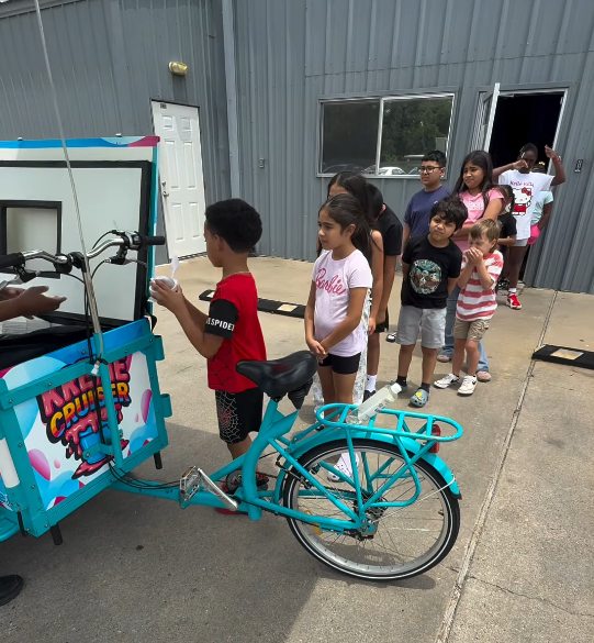 A line of children waiting at the Kreme Cruiser cart outside a building while a server hands out a cup