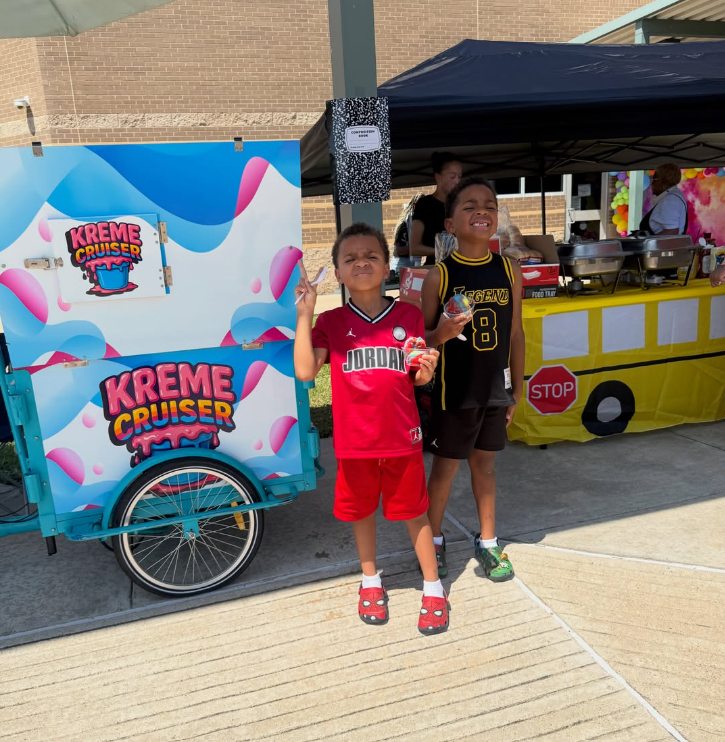 Two boys holding cups of water ice in front of the Kreme Cruiser cart at a school event